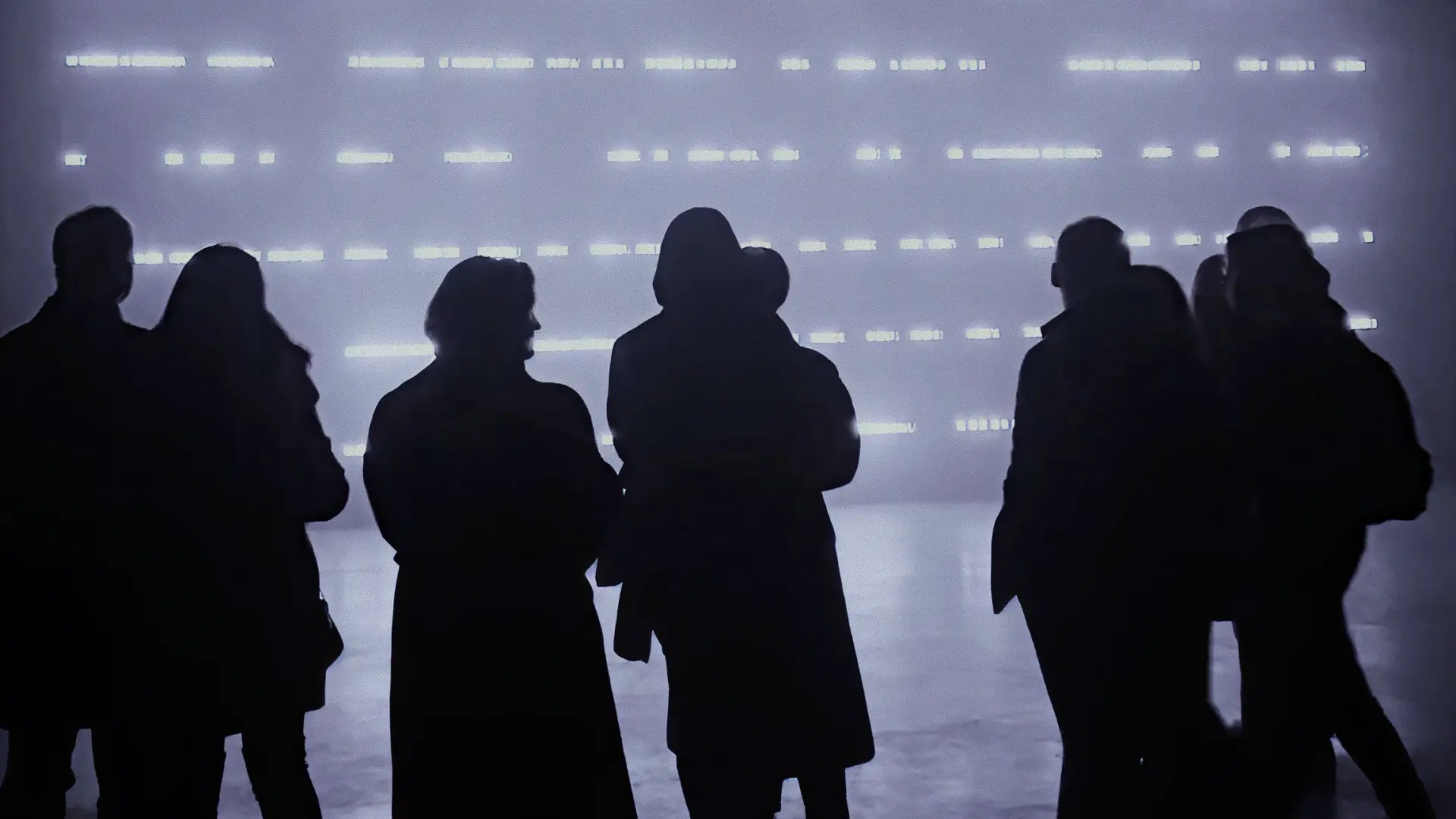 Silhouettes of people standing and facing a large illuminated digital screen with horizontal lines of light.
